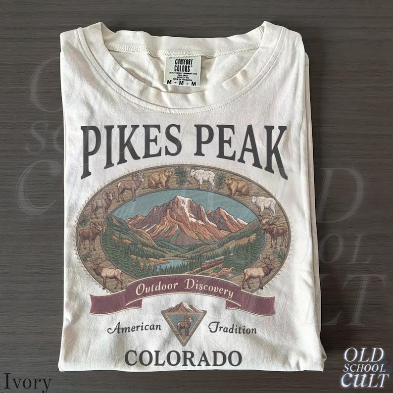 May include: Ivory-colored t-shirt featuring a graphic of Pikes Peak, Colorado. The design includes an oval illustration of the mountain, wildlife, and the text "Outdoor Discovery", "American Tradition", and "Colorado".