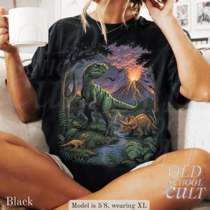 Op de afbeelding: Zwart T-shirt met een dinosaurussc&egrave;ne, waaronder een Tyrannosaurus Rex, Triceratops en een vulkaan. Het shirt heeft de tekst "OLD SCHOOL CULT". Het model is 173 cm lang en draagt XL.