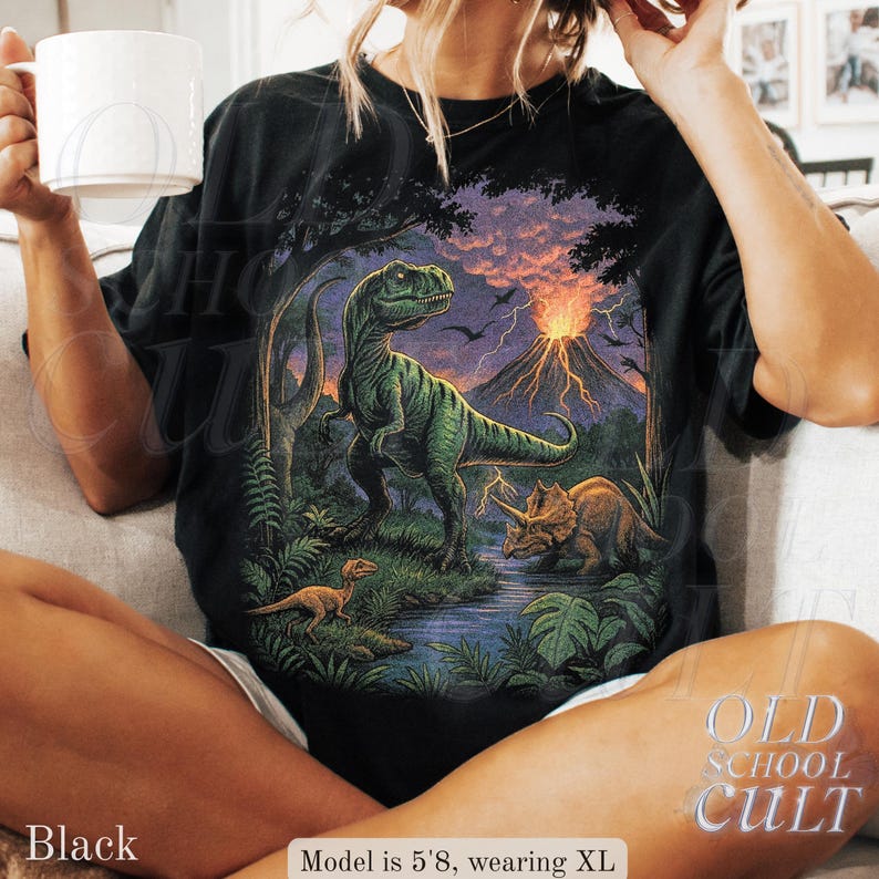 Op de afbeelding: Zwart T-shirt met een dinosaurussc&egrave;ne, waaronder een Tyrannosaurus Rex, Triceratops en een vulkaan. Het shirt heeft de tekst "OLD SCHOOL CULT". Het model is 173 cm lang en draagt XL.