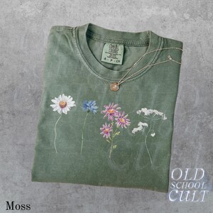 Pu&ograve; includere: Una maglietta verde muschio con un design floreale e una collana d'oro. La maglietta presenta una margherita, un fiore blu, fiori rosa e fiori bianchi. Il testo "OLD SCHOOL CULT" &egrave; visibile in basso a destra. La maglietta ha un'etichetta Comfort Colors.