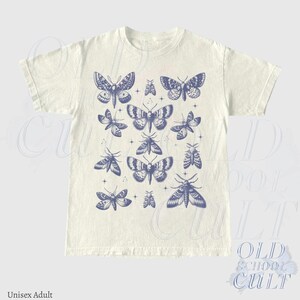 May include: Cream-colored unisex adult t-shirt featuring a collection of blue moth illustrations. The moths vary in size and design, creating a visually appealing pattern. The shirt has a classic crew neck and short sleeves.