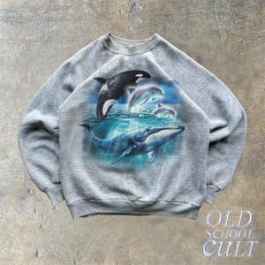 May include: A gray sweatshirt with a graphic print of a whale and dolphins swimming in the ocean. The graphic is in shades of blue and white.