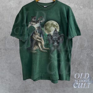 K&ouml;nnte beinhalten: Gr&uuml;nes T-Shirt mit einem Grafikdruck von drei Werw&ouml;lfen, die zum Mond heulen. Die Werw&ouml;lfe sind in einem realistischen Stil mit detailliertem Fell und scharfen Z&auml;hnen dargestellt.