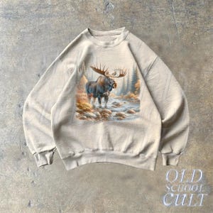 May include: A beige sweatshirt with a graphic print of a moose standing in a forest scene. The moose is brown with large antlers. The scene is surrounded by trees and a river.