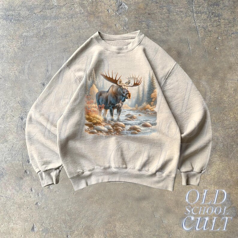 May include: A beige sweatshirt with a graphic print of a moose standing in a forest scene. The moose is brown with large antlers. The scene is surrounded by trees and a river.