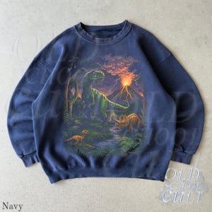 Op de afbeelding: Marineblauwe sweatshirt met een kleurrijke dinosaurussc&egrave;ne, inclusief een T-Rex, Triceratops en een uitbarstende vulkaan. De afbeelding heeft een vintage stijl. Het woord "Navy" staat linksonder.