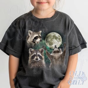 May include: Dark gray t-shirt featuring a whimsical design of three raccoons and a full moon. One raccoon is howling, another is looking up, and the third is looking forward. The text "OLD SCHOOL CULT" is printed on the bottom right.