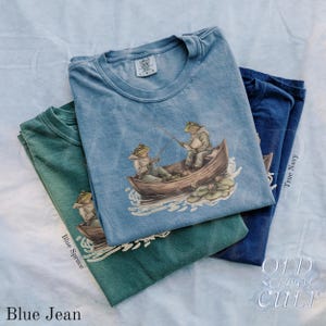 Pode incluir: Tr&ecirc;s camisetas em azul, verde e marinho est&atilde;o empilhadas. Cada camisa apresenta um gr&aacute;fico de dois sapos pescando em um barco. Os textos "Blue Jean", "Blue Spruce" e "True Navy" s&atilde;o impressos nas camisas. A marca "OLD SCHOOL CULT" tamb&eacute;m &eacute; vis&iacute;vel.