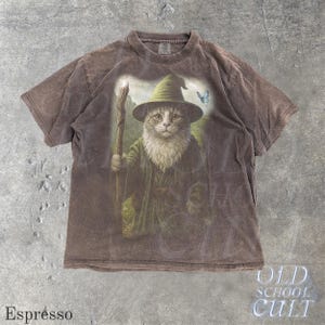 May include: Espresso-colored t-shirt featuring a whimsical illustration of a cat dressed as a wizard, complete with a hat, staff, and a butterfly. The shirt has a vintage, faded look, with the words "OLD SCHOOL CULT" printed on the bottom right.