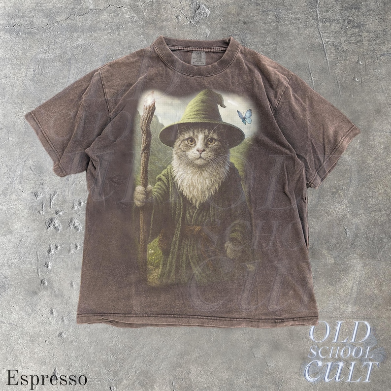 May include: Espresso-colored t-shirt featuring a whimsical illustration of a cat dressed as a wizard, complete with a hat, staff, and a butterfly. The shirt has a vintage, faded look, with the words "OLD SCHOOL CULT" printed on the bottom right.