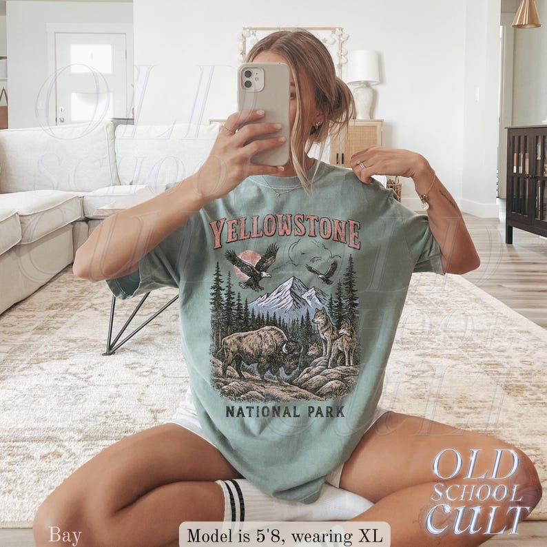 May include: A light green t-shirt with a graphic of Yellowstone National Park. The design includes a bison, wolves, mountains, and eagles. The text "YELLOWSTONE NATIONAL PARK" is printed across the top and bottom of the graphic.