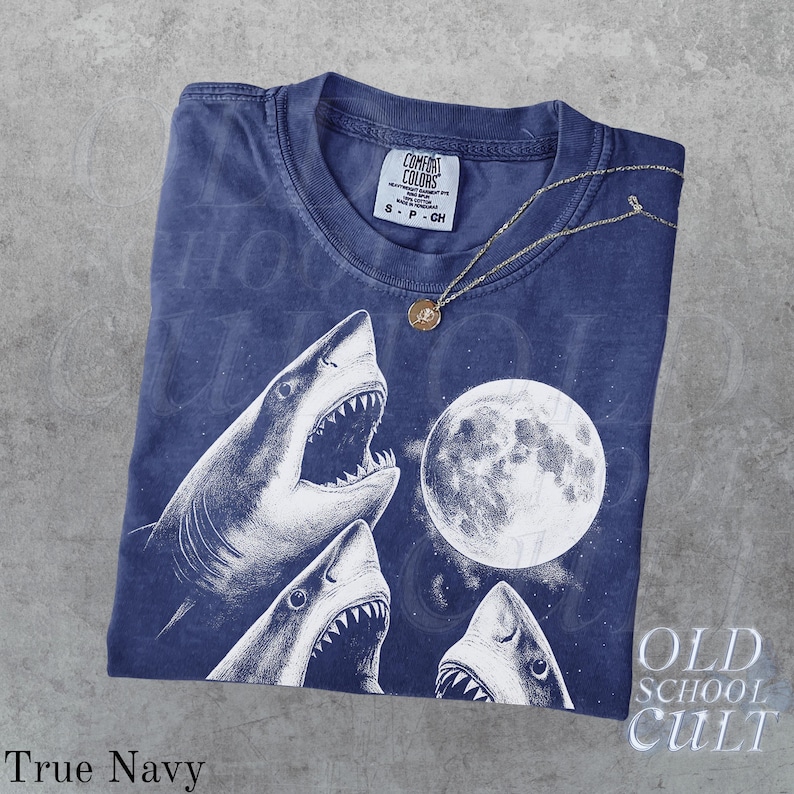 May include: True Navy t-shirt with a graphic of three sharks with open mouths and a full moon. The shirt has the text "OLD SCHOOL CULT" and "COMFORT COLORS" on the tag. A gold necklace with a pendant is on the shirt.
