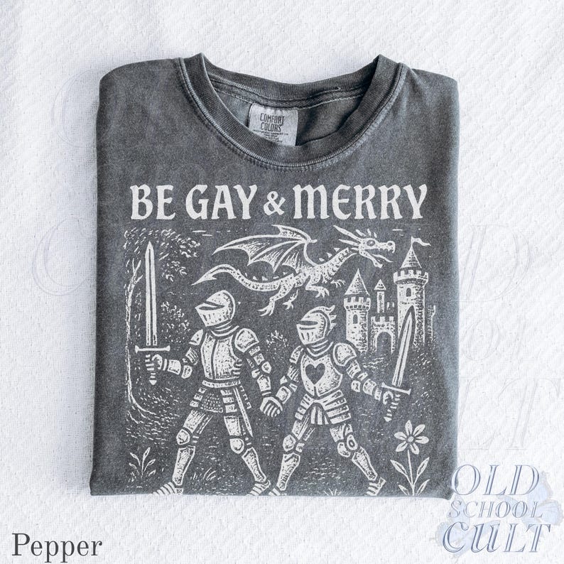 Puede incluir: Camiseta color pimienta con el texto "BE GAY & MERRY" sobre un gr&aacute;fico blanco de dos caballeros, un drag&oacute;n y un castillo. El dise&ntilde;o tiene un aspecto vintage y desgastado. La camiseta tiene cuello redondo y mangas cortas.