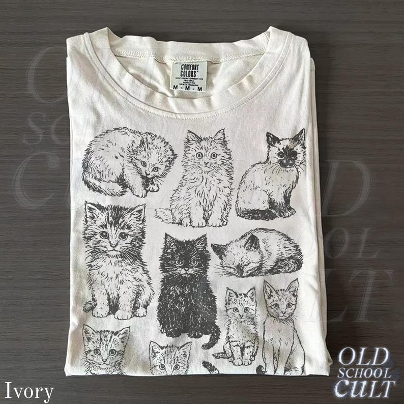 May include: Ivory-colored t-shirt featuring a collection of illustrated cats in various poses. The graphic includes kittens and adult cats in black and white. The shirt has a crew neck and the brand "Comfort Colors" is visible.