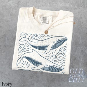 May include: Ivory-colored t-shirt with a graphic of two whales swimming in the ocean. The whales are dark blue, and the shirt has the words "Old School Cult" printed on the bottom right. A gold necklace with a pendant is draped over the shirt.