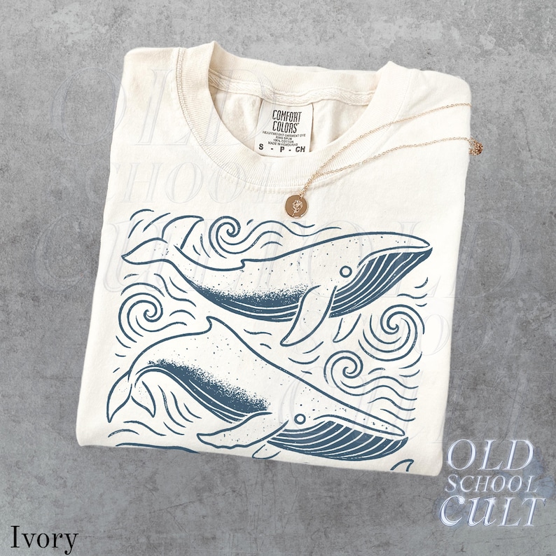 May include: Ivory-colored t-shirt with a graphic of two whales swimming in the ocean. The whales are dark blue, and the shirt has the words "Old School Cult" printed on the bottom right. A gold necklace with a pendant is draped over the shirt.