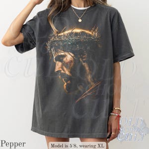May include: A dark gray t-shirt featuring a detailed portrait of a man with a crown of thorns. The image is a side profile with a warm color palette. The text "OLD SCHOOL CULT" is visible in the background.