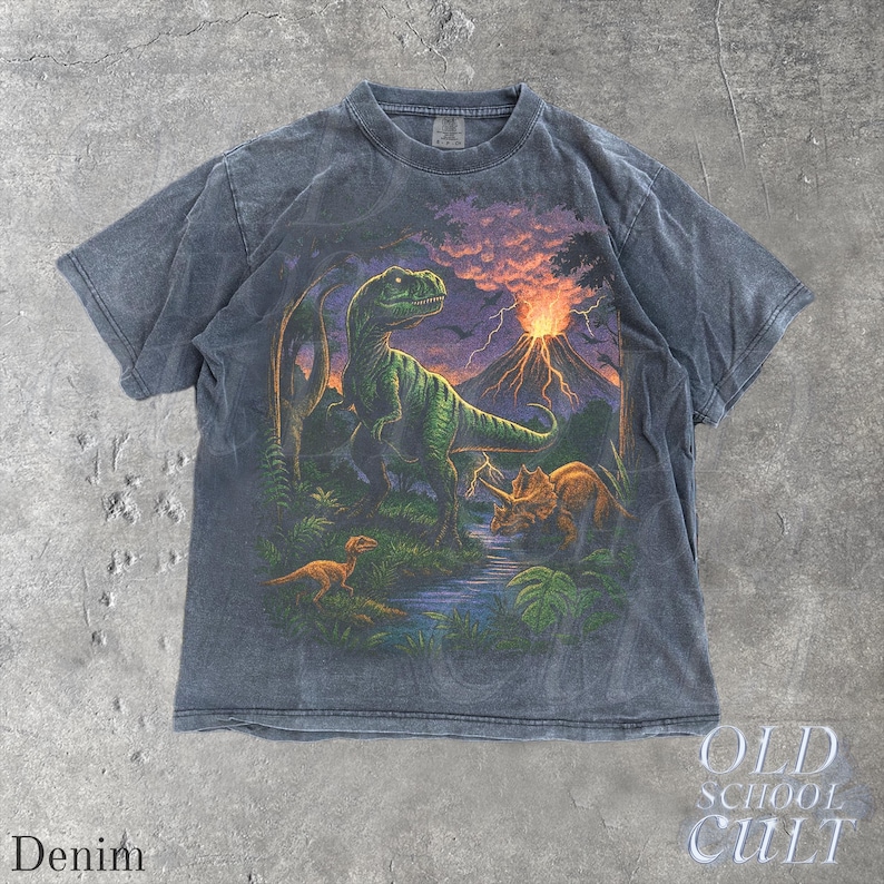 Op de afbeelding: Denimkleurig T-shirt met een levendige prehistorische sc&egrave;ne met dinosaurussen, een vulkaan en weelderig gebladerte. De afbeelding toont een groene Tyrannosaurus Rex, een Triceratops en andere dinosaurussen. Het shirt heeft de tekst "OLD SCHOOL CULT" en "Denim".