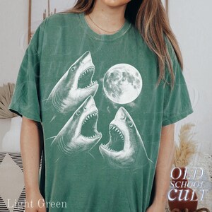 May include: Light green t-shirt featuring a graphic of three white sharks with open mouths and a full moon. The text "OLD SCHOOL CULT" is printed on the lower right side of the shirt. The words "Light Green" are printed on the lower left side.