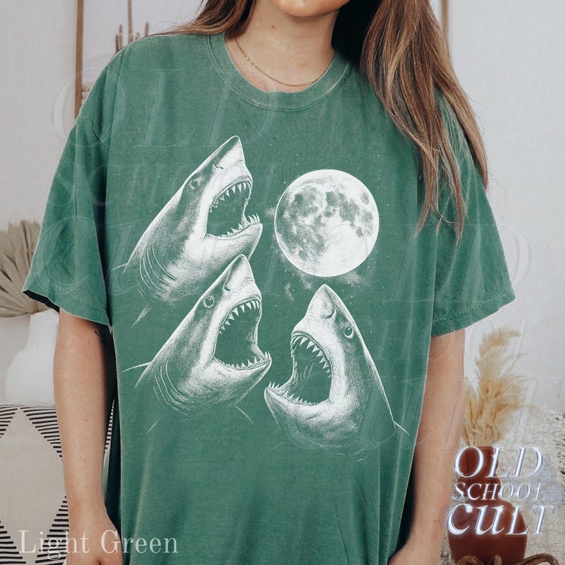 May include: Light green t-shirt featuring a graphic of three white sharks with open mouths and a full moon. The text "OLD SCHOOL CULT" is printed on the lower right side of the shirt. The words "Light Green" are printed on the lower left side.