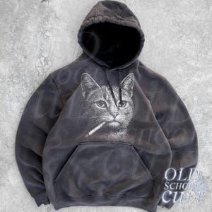 Könnte beinhalten: Dunkelgrauer Kapuzenpullover mit einem Aufdruck einer Katze mit Zigarette. Der Hoodie hat eine Kapuze mit Kordelzug und eine Kängurutasche. Der Text "OLD SCHOOL CULT" ist unten rechts aufgedruckt.