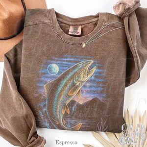 May include: Espresso-coloured sweatshirt with a large fish leaping from water, a moon, and mountains. The crew neck sweatshirt has long sleeves. A silver necklace with a pendant is draped on the shirt. The text "Espresso" is at the bottom.