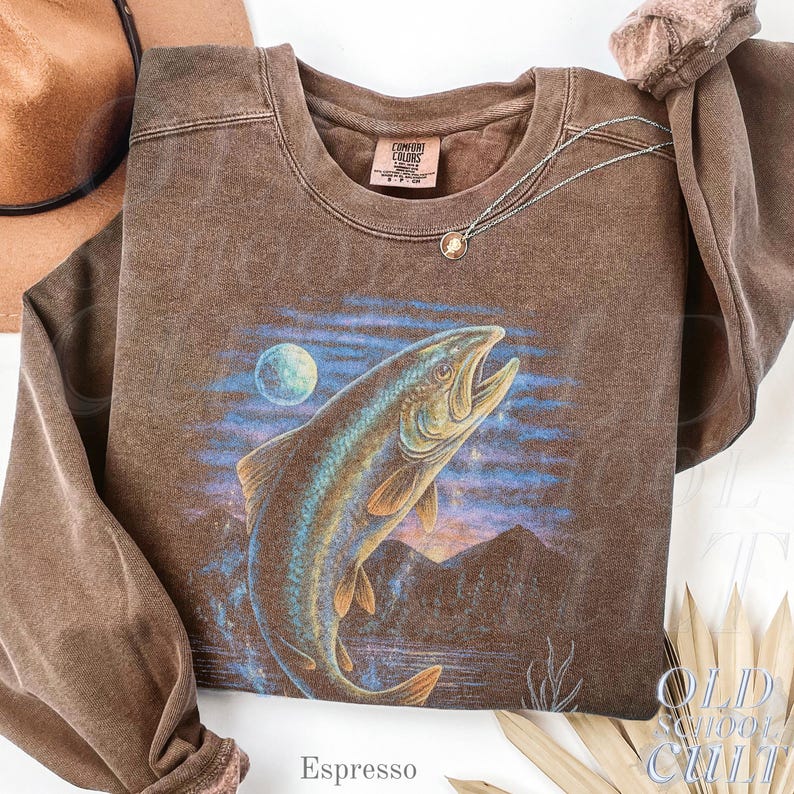 May include: Espresso-coloured sweatshirt with a large fish leaping from water, a moon, and mountains. The crew neck sweatshirt has long sleeves. A silver necklace with a pendant is draped on the shirt. The text "Espresso" is at the bottom.