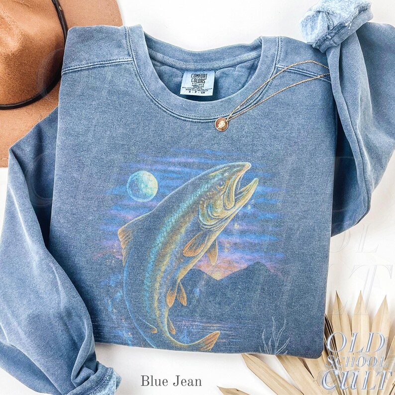 May include: Blue jean-coloured sweatshirt with a large illustration of a jumping fish, moon, and mountain landscape. The crew neck sweatshirt has long sleeves. The text "Blue Jean" is at the bottom.