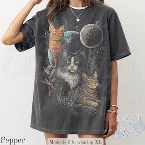 以下が含まれることがあります： 宇宙をテーマにしたデザインの、3匹の猫、惑星、星が描かれた濃いグレーのTシャツ。猫はオレンジ、白と黒、茶色とタン色です。このシャツはヴィンテージ感があります。