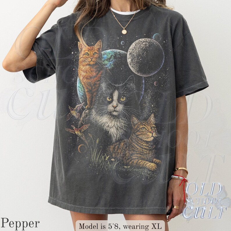 以下が含まれることがあります： 宇宙をテーマにしたデザインの、3匹の猫、惑星、星が描かれた濃いグレーのTシャツ。猫はオレンジ、白と黒、茶色とタン色です。このシャツはヴィンテージ感があります。