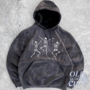 Puede incluir: Sudadera con capucha gris oscuro con bolsillo delantero y cordón. La sudadera presenta un gráfico blanco de tres esqueletos bailando. El texto "OLD SCHOOL CULT" está impreso en la parte inferior derecha.