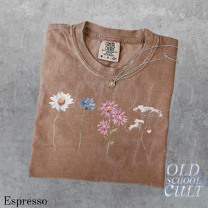 Pu&ograve; includere: T-shirt color espresso con girocollo e maniche corte. La maglietta presenta un design floreale con una margherita, un fiordaliso e altri fiori. Una collana d'argento con un ciondolo poggia sulla maglietta. La maglietta ha il testo "OLD SCHOOL CULT".