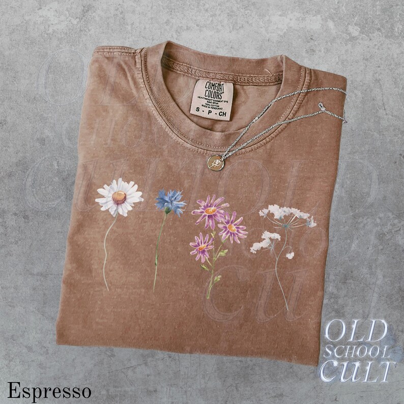 Pu&ograve; includere: T-shirt color espresso con girocollo e maniche corte. La maglietta presenta un design floreale con una margherita, un fiordaliso e altri fiori. Una collana d'argento con un ciondolo poggia sulla maglietta. La maglietta ha il testo "OLD SCHOOL CULT".