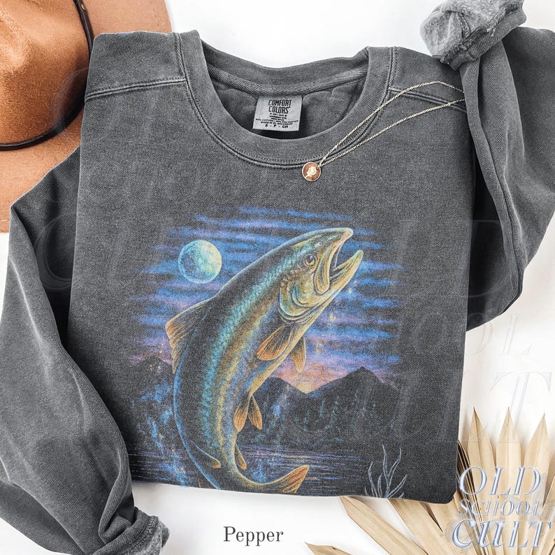 May include: Dark grey sweatshirt with a leaping fish graphic, moon, and mountain scene. The crew neck sweatshirt has long sleeves. The brand tag is visible on the neckline. The text "OLD SCHOOL CULT" is printed on the bottom right.