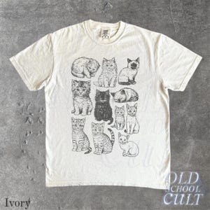 May include: Ivory-colored t-shirt featuring a collection of illustrated cats in various poses. The graphic is in black and white, with the words "OLD SCHOOL CULT" printed in a vintage style. The word "Ivory" is printed at the bottom left.