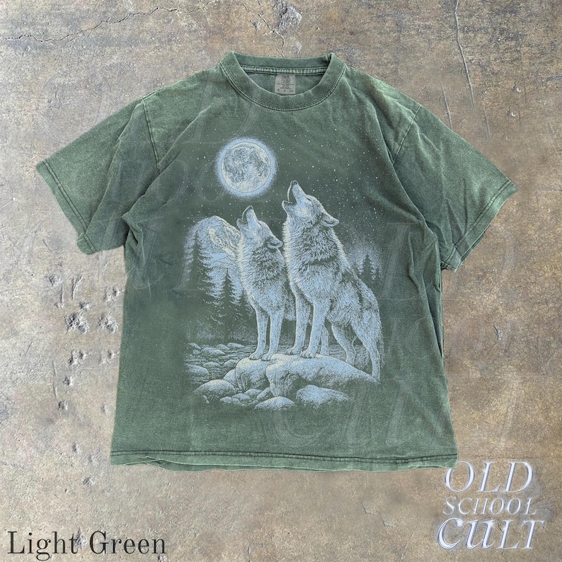 May include: Light green t-shirt featuring a graphic of two wolves howling at a full moon. The design includes a mountain range and forest. The shirt has the text "OLD SCHOOL CULT" and "Light Green".