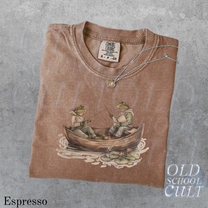 Pode incluir: Camiseta cor espresso com um gr&aacute;fico de dois sapos pescando em um barco. A camiseta tem gola redonda e mangas curtas. Um colar de prata com um pequeno pingente repousa sobre a camiseta. A etiqueta da marca diz "Comfort Colors". O texto "Old School Cult" &eacute; vis&iacute;vel.