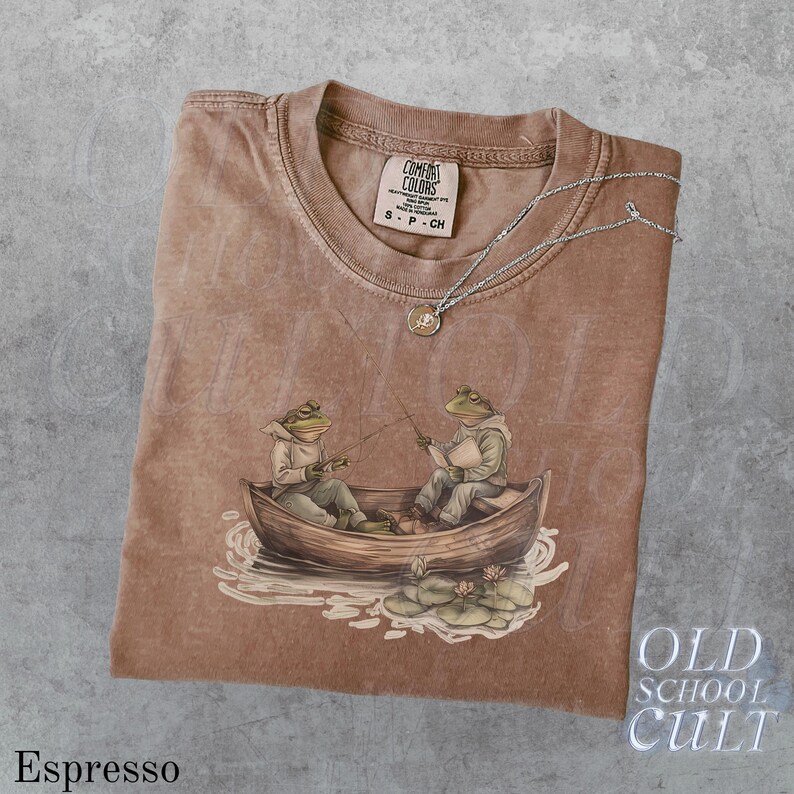 Pode incluir: Camiseta cor espresso com um gr&aacute;fico de dois sapos pescando em um barco. A camiseta tem gola redonda e mangas curtas. Um colar de prata com um pequeno pingente repousa sobre a camiseta. A etiqueta da marca diz "Comfort Colors". O texto "Old School Cult" &eacute; vis&iacute;vel.