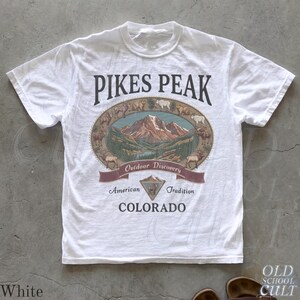 May include: White t-shirt with a graphic featuring "PIKES PEAK" in black lettering above an oval illustration of a mountain landscape. The shirt also includes the words "Outdoor Discovery," "American Tradition," and "COLORADO."