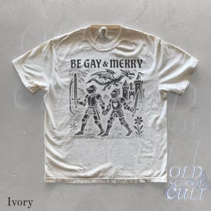 Puede incluir: Camiseta de color marfil con el texto "BE GAY & MERRY" sobre una ilustraci&oacute;n negra de dos caballeros, un drag&oacute;n y un castillo. El dise&ntilde;o es de inspiraci&oacute;n vintage y tiene un tema medieval.