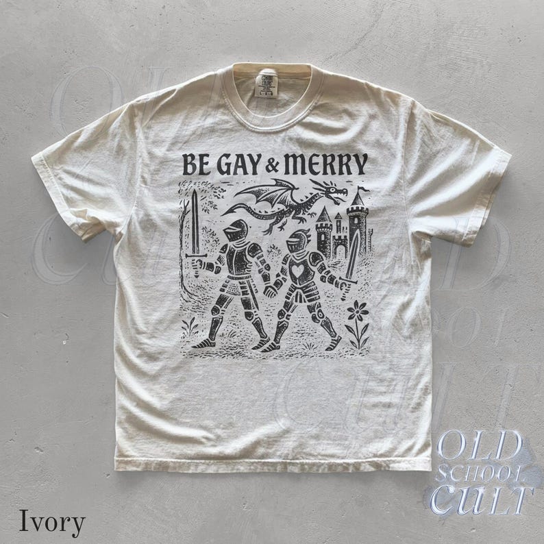 Puede incluir: Camiseta de color marfil con el texto "BE GAY & MERRY" sobre una ilustraci&oacute;n negra de dos caballeros, un drag&oacute;n y un castillo. El dise&ntilde;o es de inspiraci&oacute;n vintage y tiene un tema medieval.