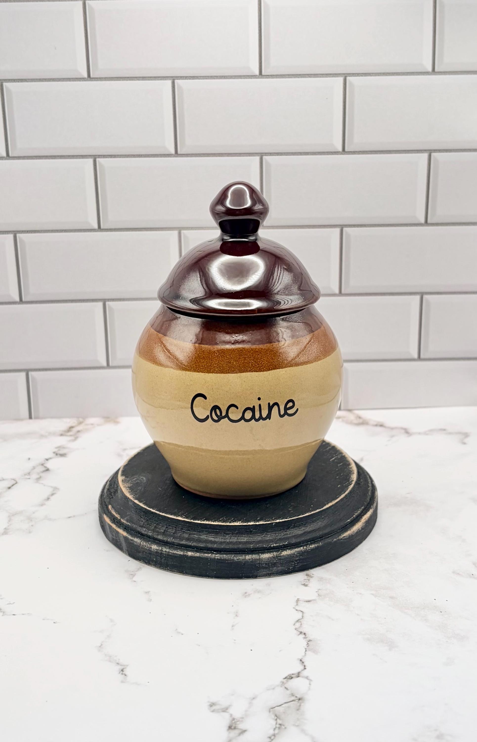 May include: A round ceramic jar with a removable lid, featuring a glossy dark brown top and a tan base. The word Cocaine is printed in black script lettering on the front of the jar. The container has a smooth, glazed finish.