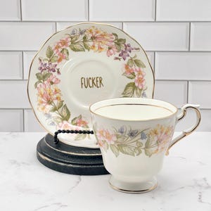 May include: A white porcelain teacup and saucer set featuring a delicate floral pattern in shades of pink, yellow, and green with gold trim. The center of the saucer displays the word FUCKER in gold lettering, matching the cup's elegant design.