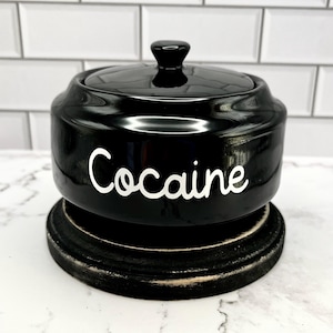 Puede incluir: Un tarro de cer&aacute;mica negro con tapa, sobre una base de madera oscura. La palabra "Cocaine" est&aacute; escrita en blanco en la parte delantera del tarro. El tarro tiene un acabado brillante.
