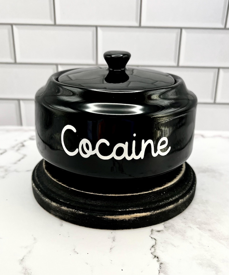 Puede incluir: Un tarro de cer&aacute;mica negro con tapa, sobre una base de madera oscura. La palabra "Cocaine" est&aacute; escrita en blanco en la parte delantera del tarro. El tarro tiene un acabado brillante.