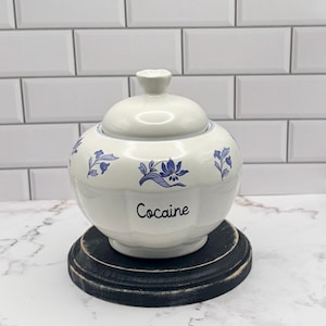 May include: A white ceramic lidded jar with a rounded, scalloped body and a matching lid. The jar features blue floral patterns painted around the exterior and the word Cocaine printed in black script lettering on the front center surface.