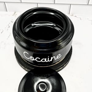 Puede incluir: Tarro de cer&aacute;mica negro con tapa, con la palabra "Cocaine" en escritura blanca. El tarro se asienta sobre una base circular oscura. La tapa est&aacute; quitada, revelando el interior del tarro.