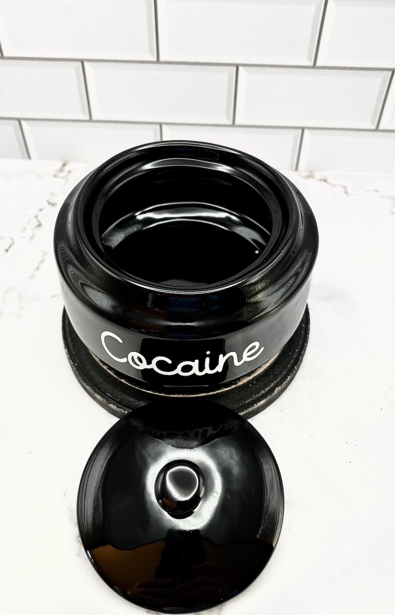Puede incluir: Tarro de cer&aacute;mica negro con tapa, con la palabra "Cocaine" en escritura blanca. El tarro se asienta sobre una base circular oscura. La tapa est&aacute; quitada, revelando el interior del tarro.