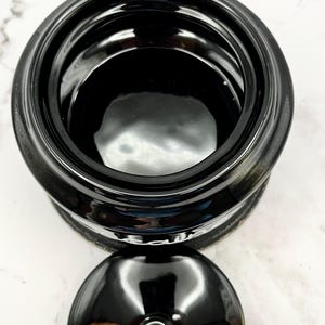 Puede incluir: Un tarro de cer&aacute;mica negro brillante con tapa a juego. El tarro tiene una abertura ancha y un cuerpo redondeado. La tapa presenta un peque&ntilde;o asa redonda. El tarro y la tapa est&aacute;n sobre una superficie blanca.
