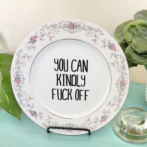 May include: White decorative plate with a floral border and the text "YOU CAN KINDLY FUCK OFF" in black. The plate is displayed on a black metal stand, with greenery and a glass paperweight in the background.